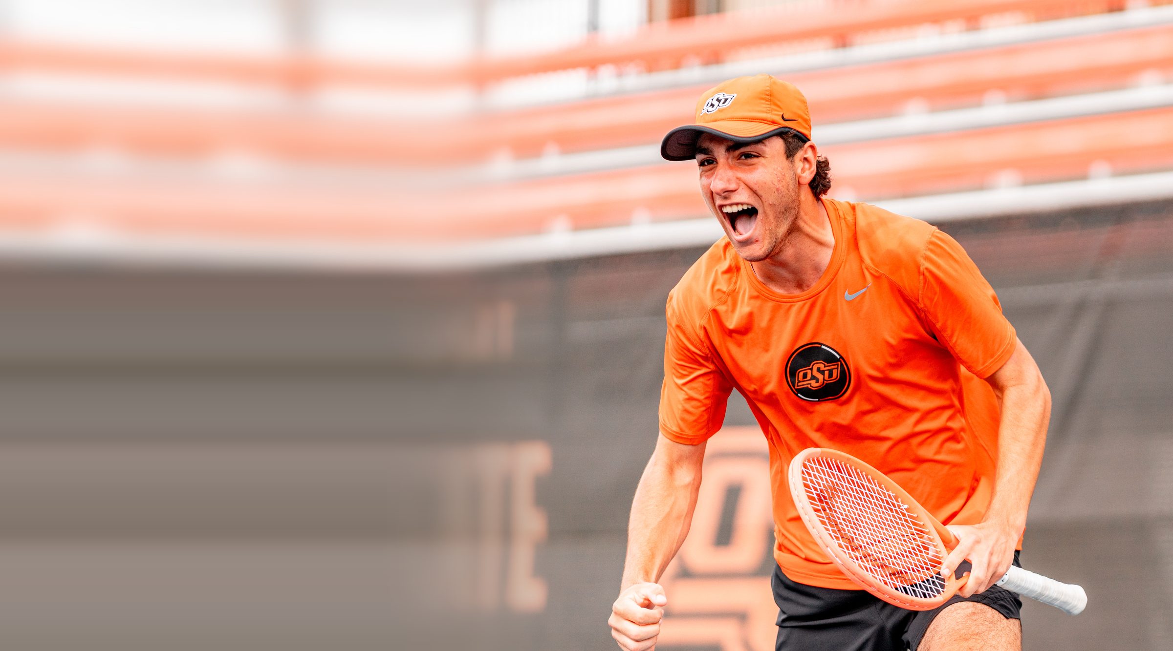 College-Tennisspieler in orangem Trikot jubelt mit geballter Faust und Schläger in der Hand